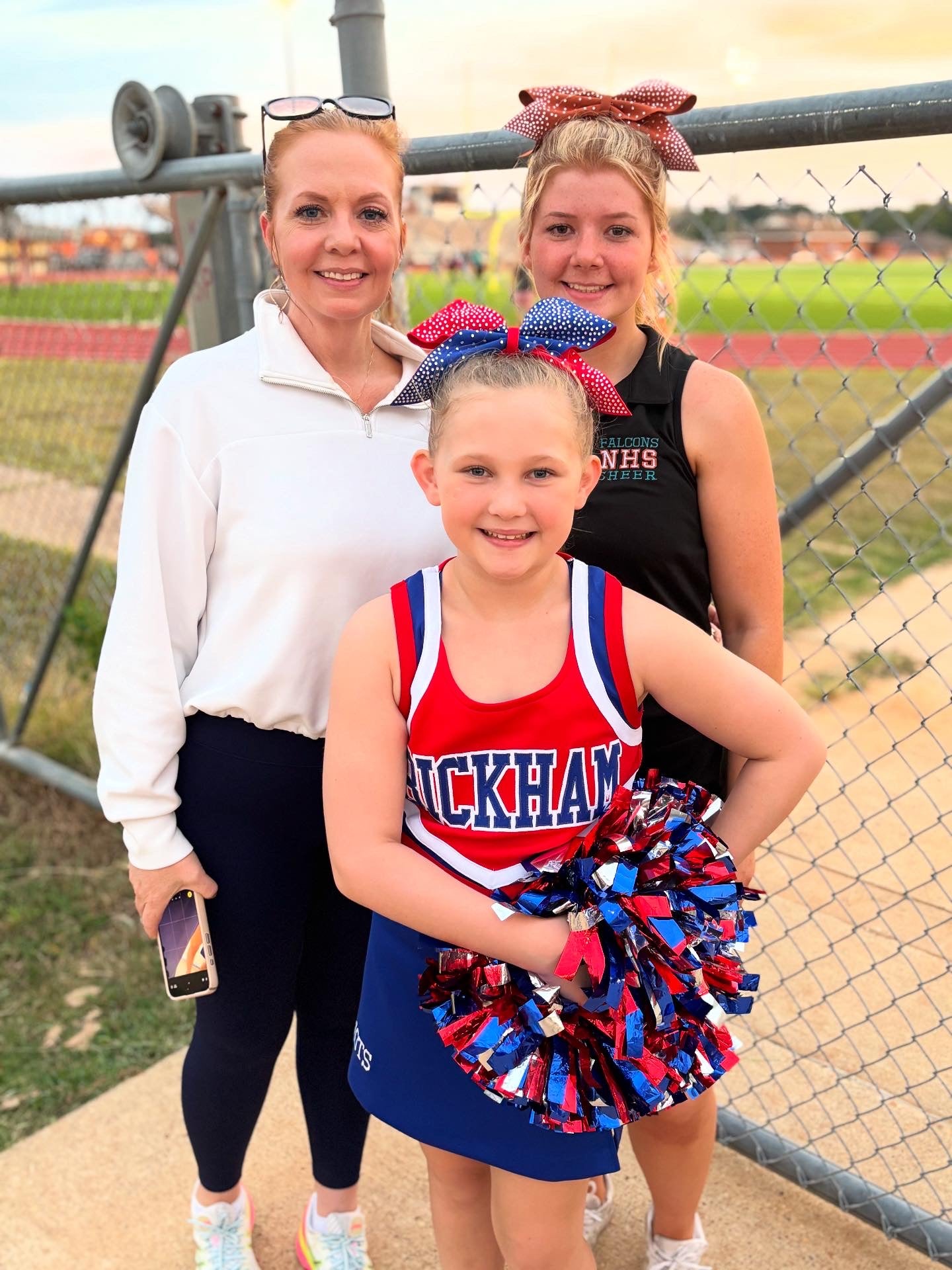Three women, including a young girl in a cheerleading outfit, standing outdoors with a sports field in the background.